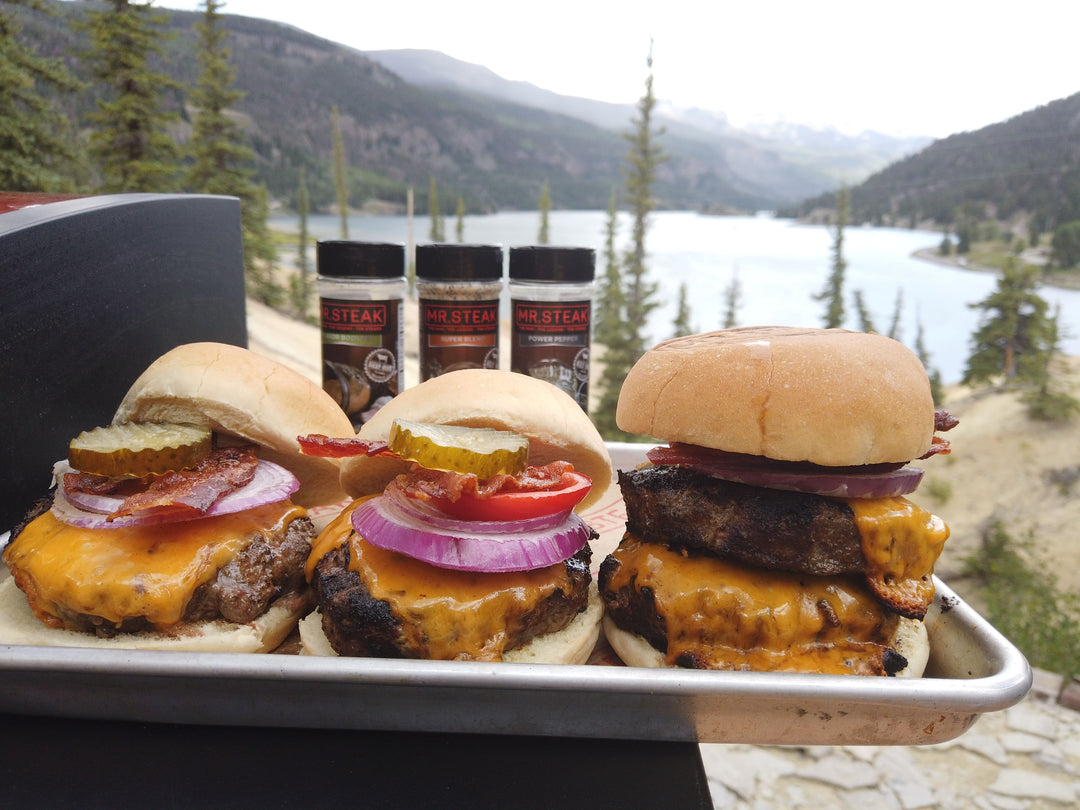 Three cheeseburgers topped with pickles, bacon, tomato, and onion with a lake in the background