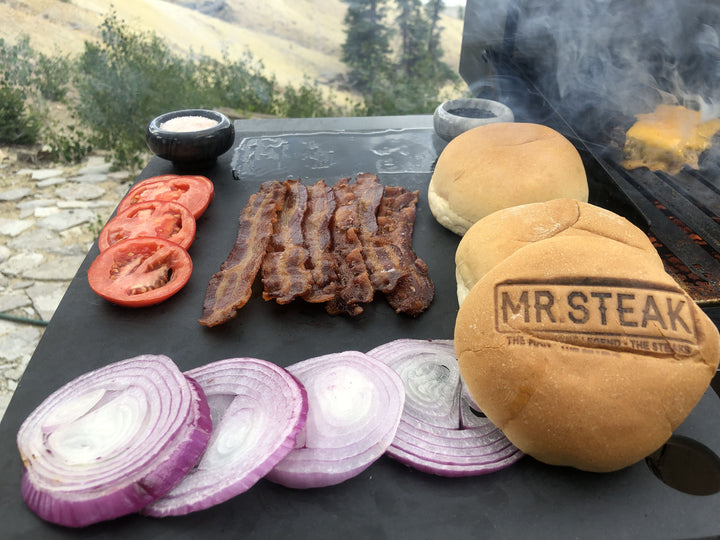 Burger grilling with onion, tomato and bacon toppings ready on the side. Mr Steak branded bun 