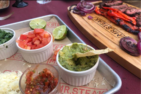 Steak fajitas ready to be made with guacamole, salsa, tomato, onion, lime, and red peppers