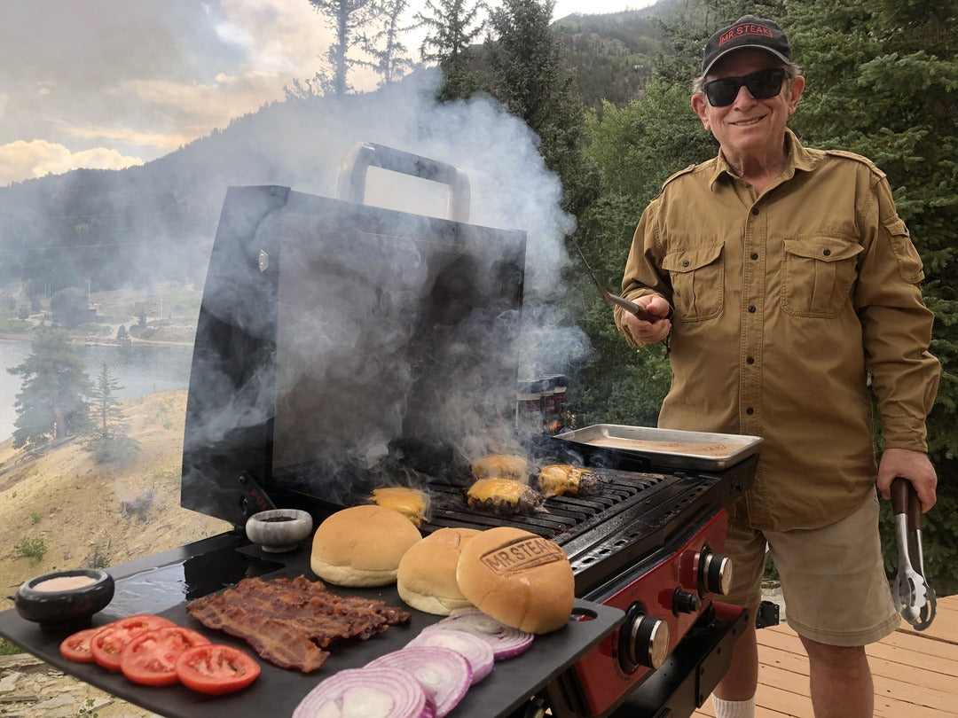 Mr. Steak grilling up some juicy burgers with cheese on top and bacon, tomato, onion and toasted buns on the side