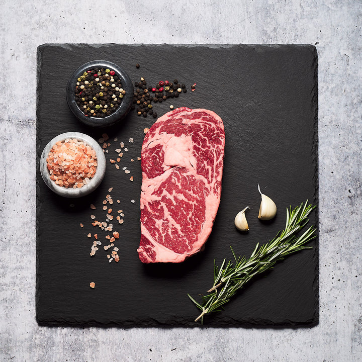 Richly marbled boneless ribeye alongside salt, pepper, garlic, and rosemary