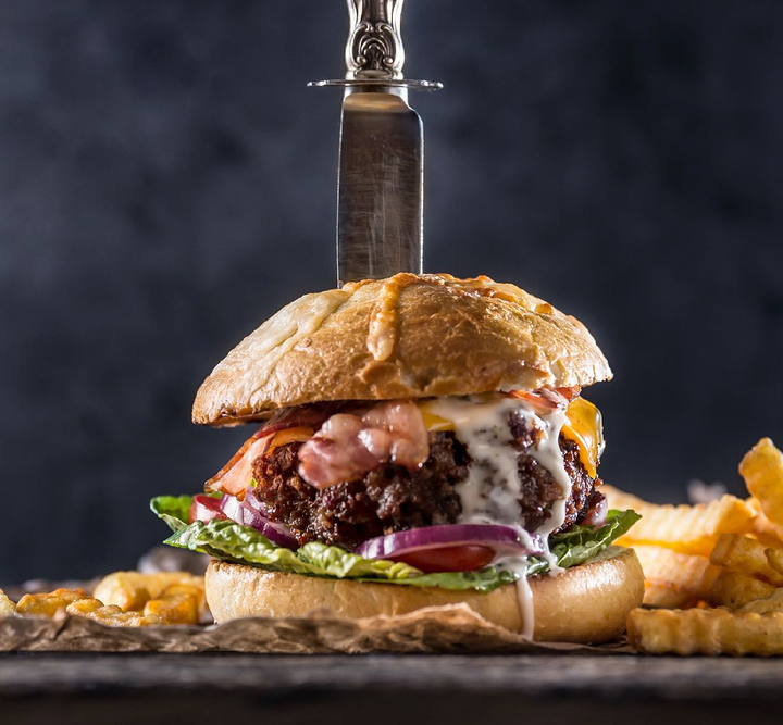 Knife going through juicy Wagyu cheeseburger dripping with sauce, topped with bacon, lettuce, and onions, on toasted cheese buns