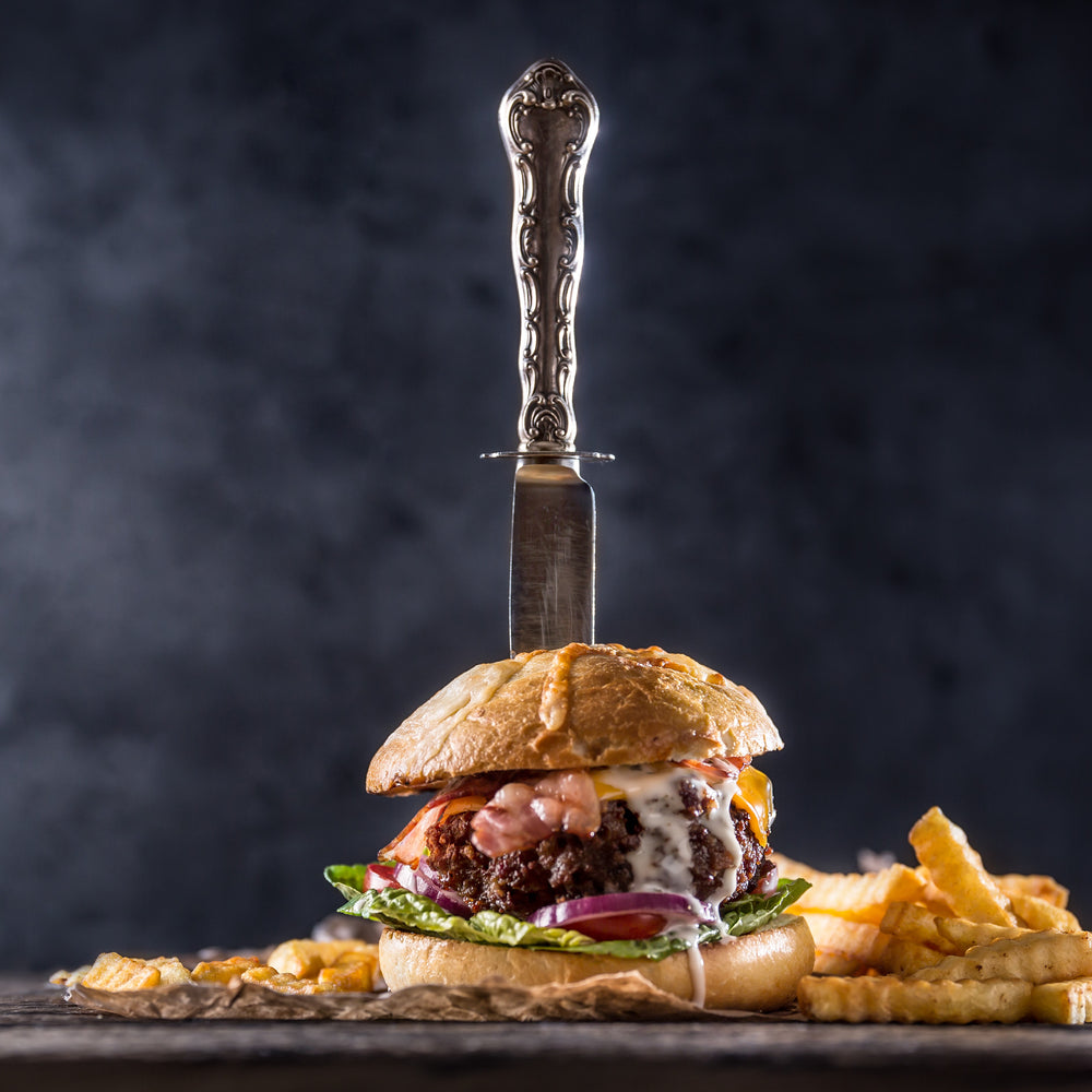 Knife going through juicy wagu burger, dripping with sauces, topped with bacon, tomato, lettuce, and cheese